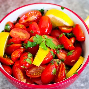 Marinated cherry tomatoes served in a red bowl. Cherry tomatoes are halved and garnished with fresh lemon slices and cilantro.
