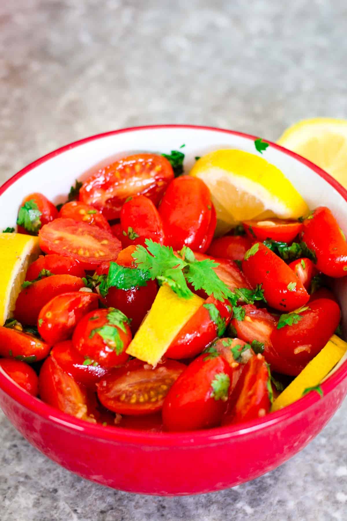Marinated Cherry Tomatoes on a red bowl, tomatoes are halved and garnished with cilantro and lemon slices.