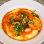 A white plate showing creamy polenta topped with shrimp, sauce and garnish.