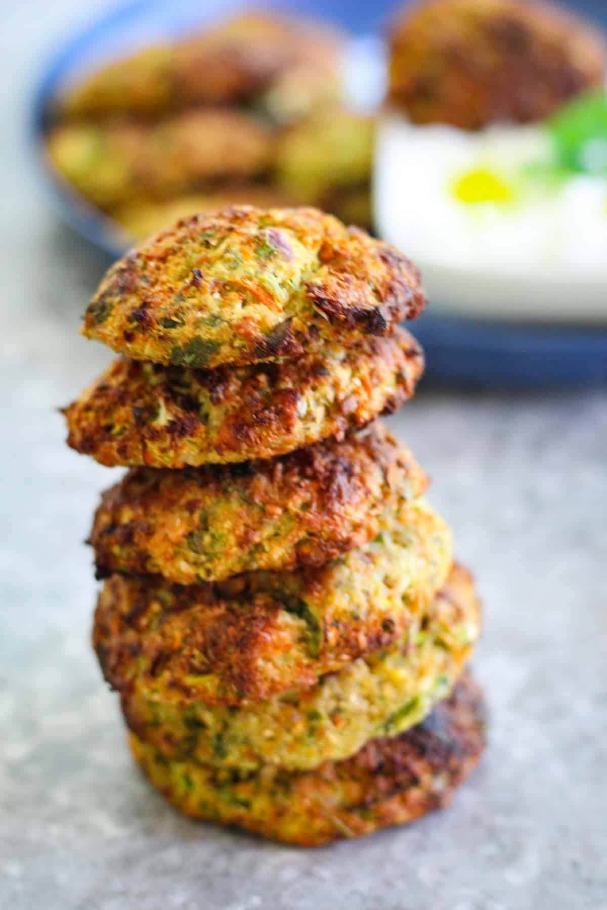 Cauliflower fritters in a tower shape over one another. In the background you can see a blurred plate with more fritters and the dip.