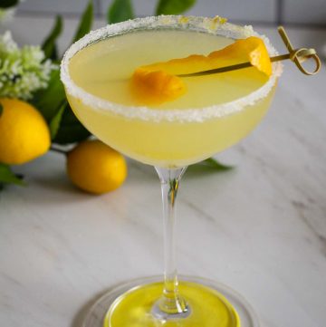 An image of a cocktail on a coaster. It's a lemon cocktail and it shows lemons and green leaves in the background.