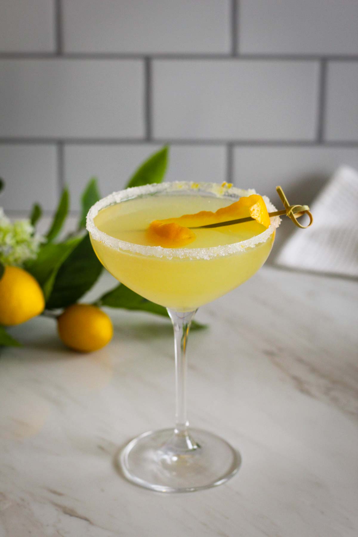 A coup cocktail glass showing a Meyer lemon drop with a peel garnish and more lemons in the background