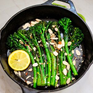 A cast iron skillet shown with sauteed broccolini, sprinkled in shaved almonds and garnished with a slice of almond.