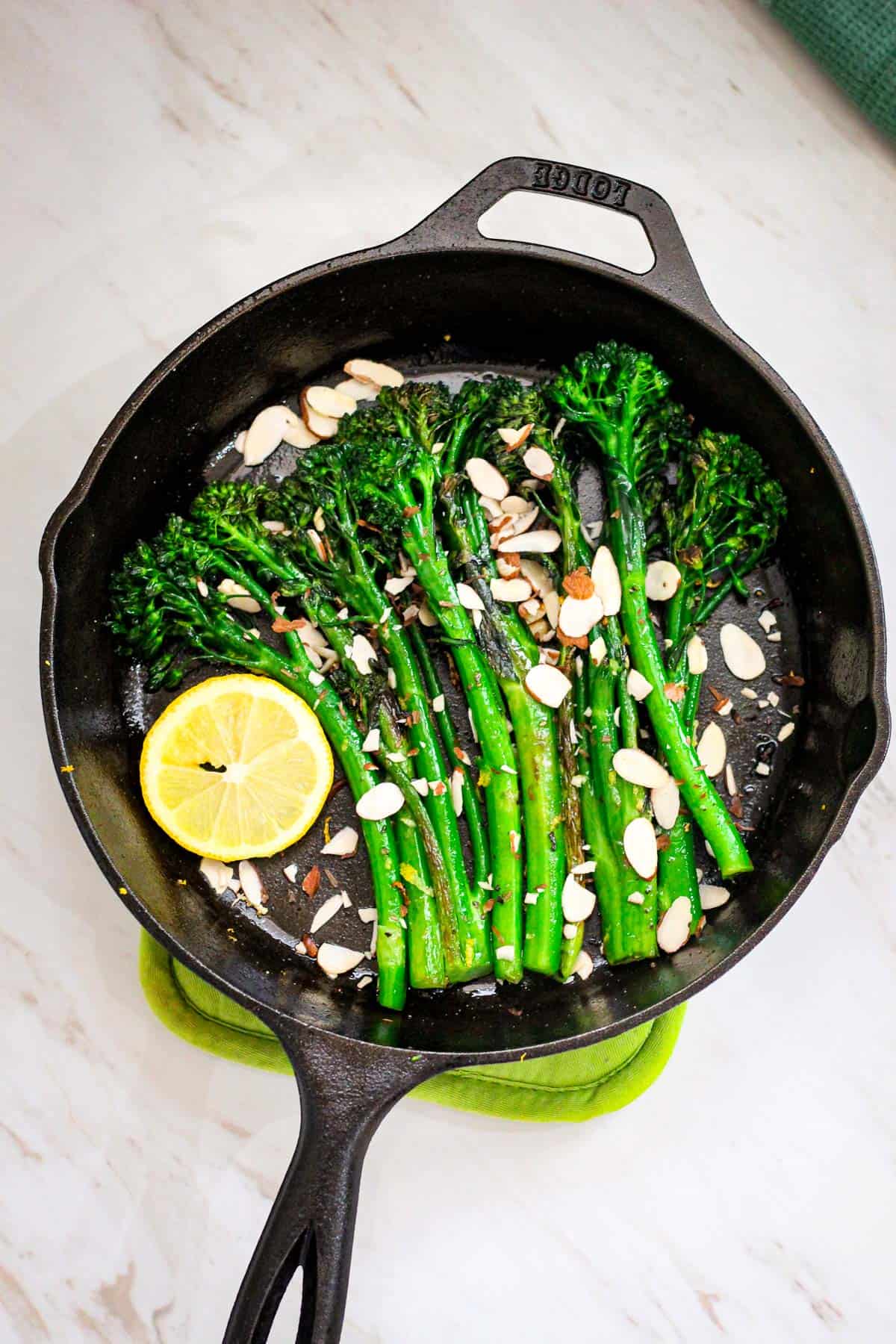 A cast iron skillet with sauteed broccolini. Skillet shows some almonds and a slice of lemon as well.