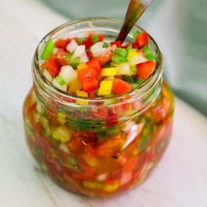 A glass jar filled to the top with salsa criolla and there's a spoon in the jar too.