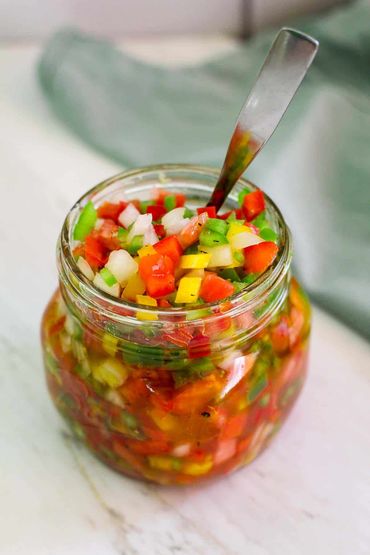 A jar of salsa made with peppers, tomatoes, onions and oil/vinegar. Jar is shown with a spoon in it, ready to serve.