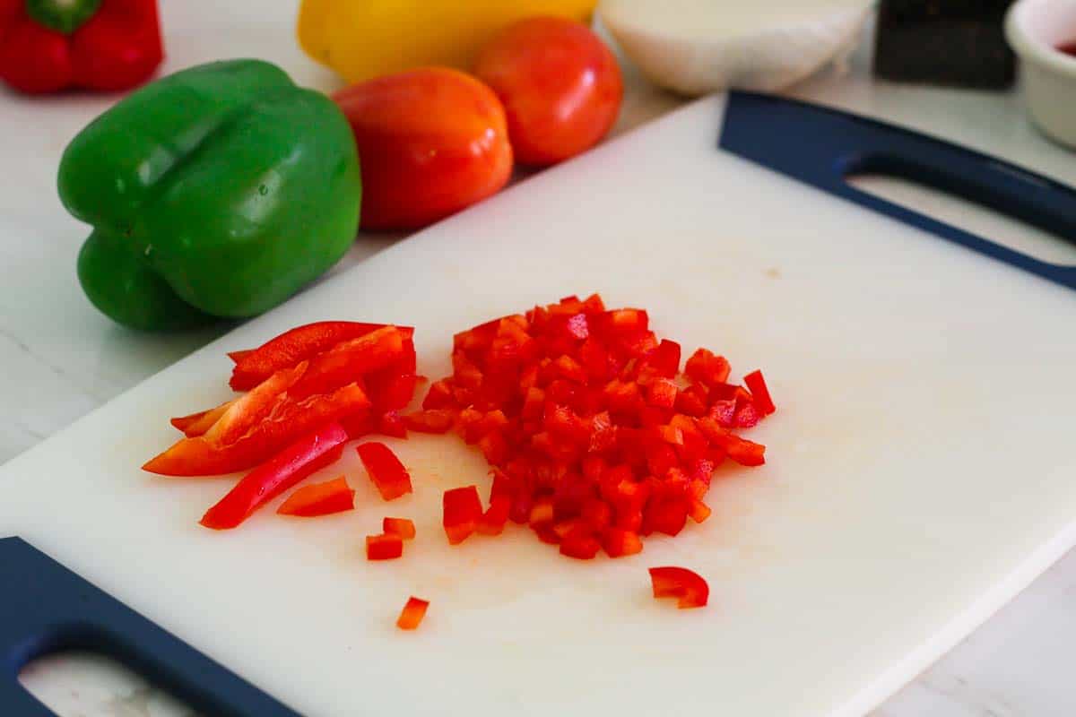 Diced red peppers on the cutting board, with the rest of the vegetables seen intact in the background.