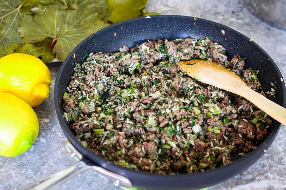 A skillet with cooked meat, rice and herbs which will make the stuffing for grape leaves. There's a wooden spoon over stuffing and 2 leamons as garnishes on the left hand side. 