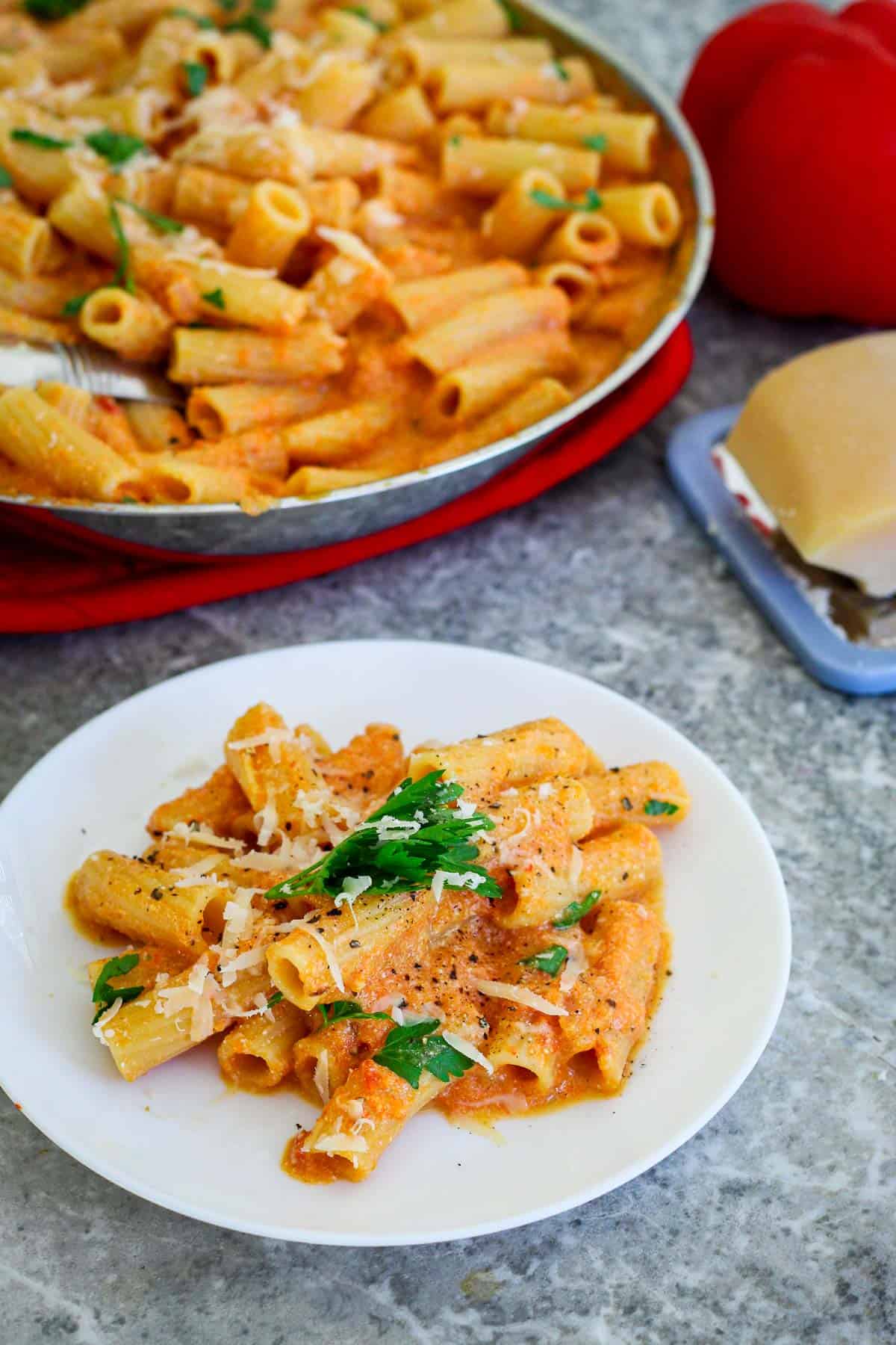 A serving of rigatoni pasta in pepper sauce. Pasta is garnished with parsley and grated cheese. Behind the plate, you can see the entire pan where pasta dish is cooked. 