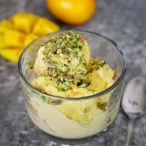A serving cup with mango gelato topped with pistachios. In the background you see a cut manga and a meyer lemon.