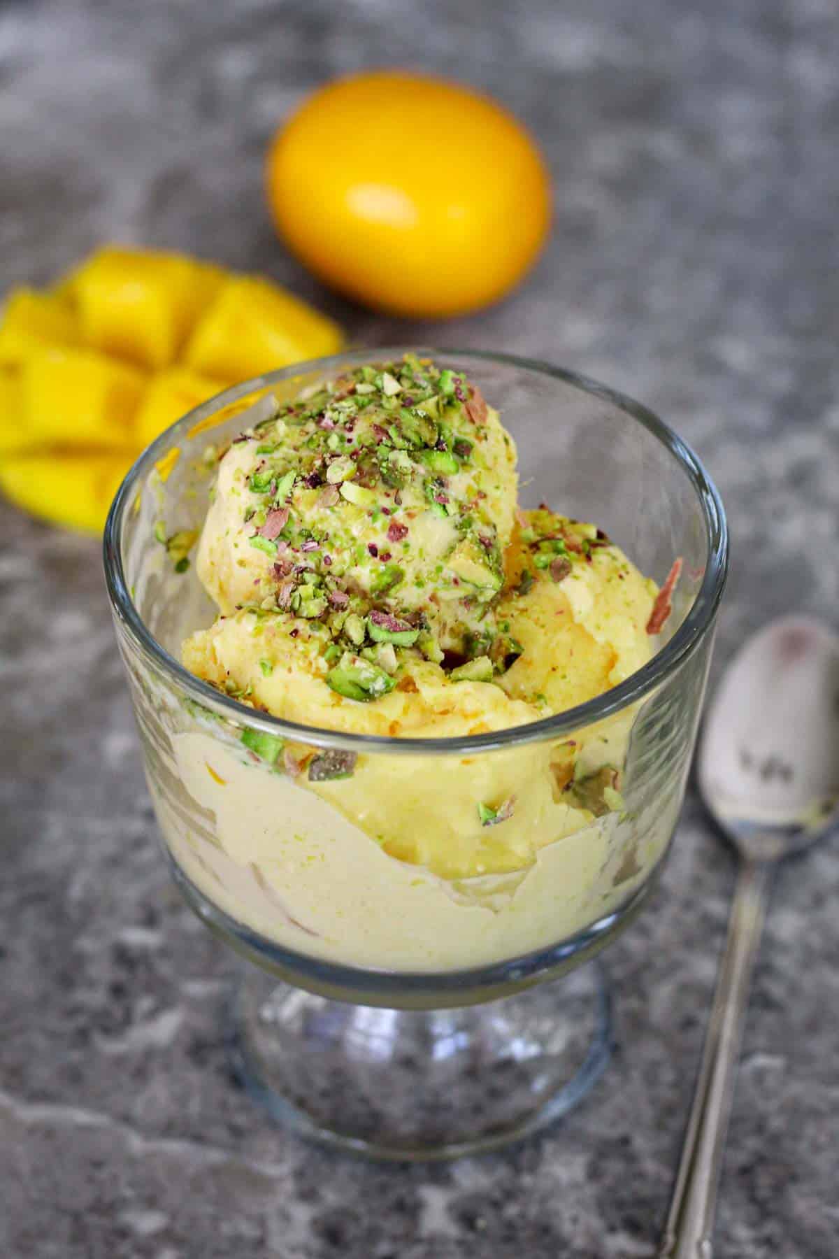 A serving of a yellow ice cream topped with chopped pistachio. In the background, there's a Meye lemon and a half mango cut in cubes.