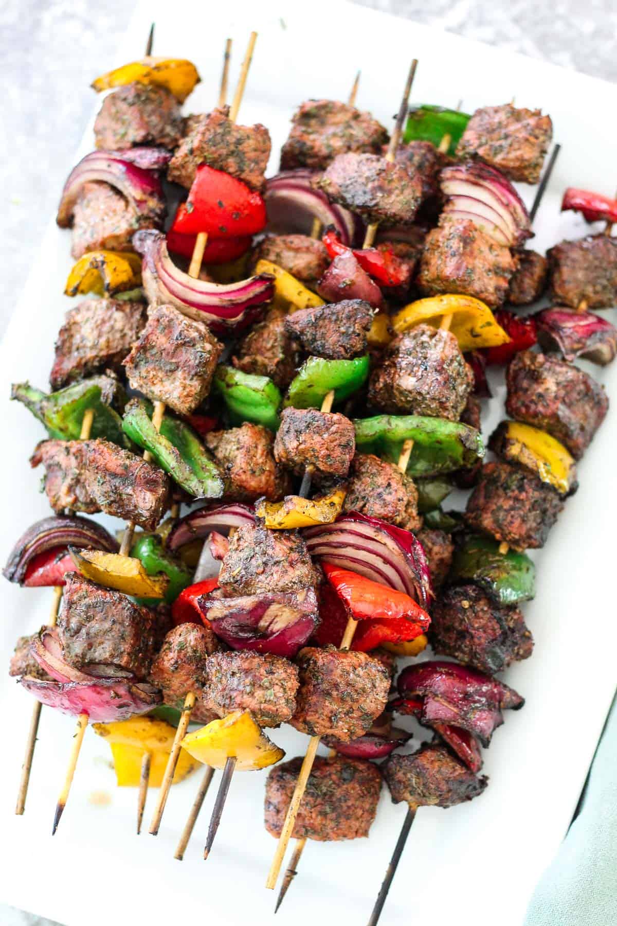 A platter with grilled beef kebabs piled on top of each other. Skewers are slightly charred on the edges and they're threaded with meat, peppers, onions.