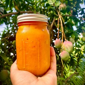 A handheld jar of mango jam in front of a mango tree with ripe mangoes.