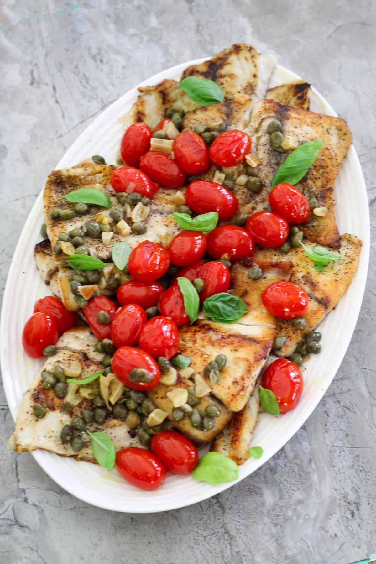 An oval white platter with white fish (barramundi) covered with capers and grape tomatoes.