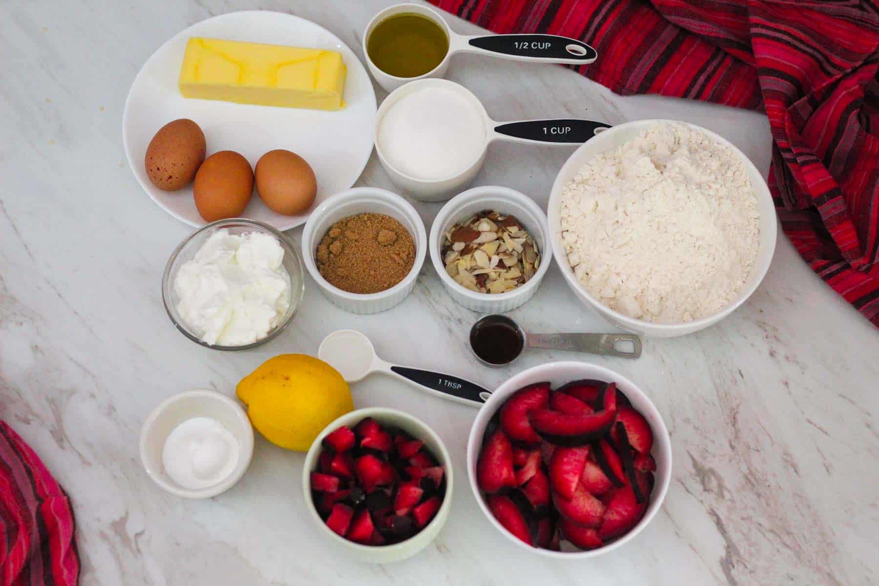 Ingredients for plum cake: butter, eggs, olive oil, yogurt, sugar, flour, almonds, brown sugar, almond extract, vanilla extract, lemon, leveners, chopped plums, sliced plums. 