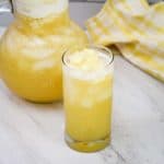 A glass of lemonade with pieces of pineapple floating on top and a pitcher of same lemonade on the back.