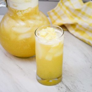 A glass of lemonade with pieces of pineapple floating on top and a pitcher of same lemonade on the back.