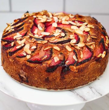 A plum almond cake, already baked but not sliced featured on a cake stand in the counter of a kitchen.