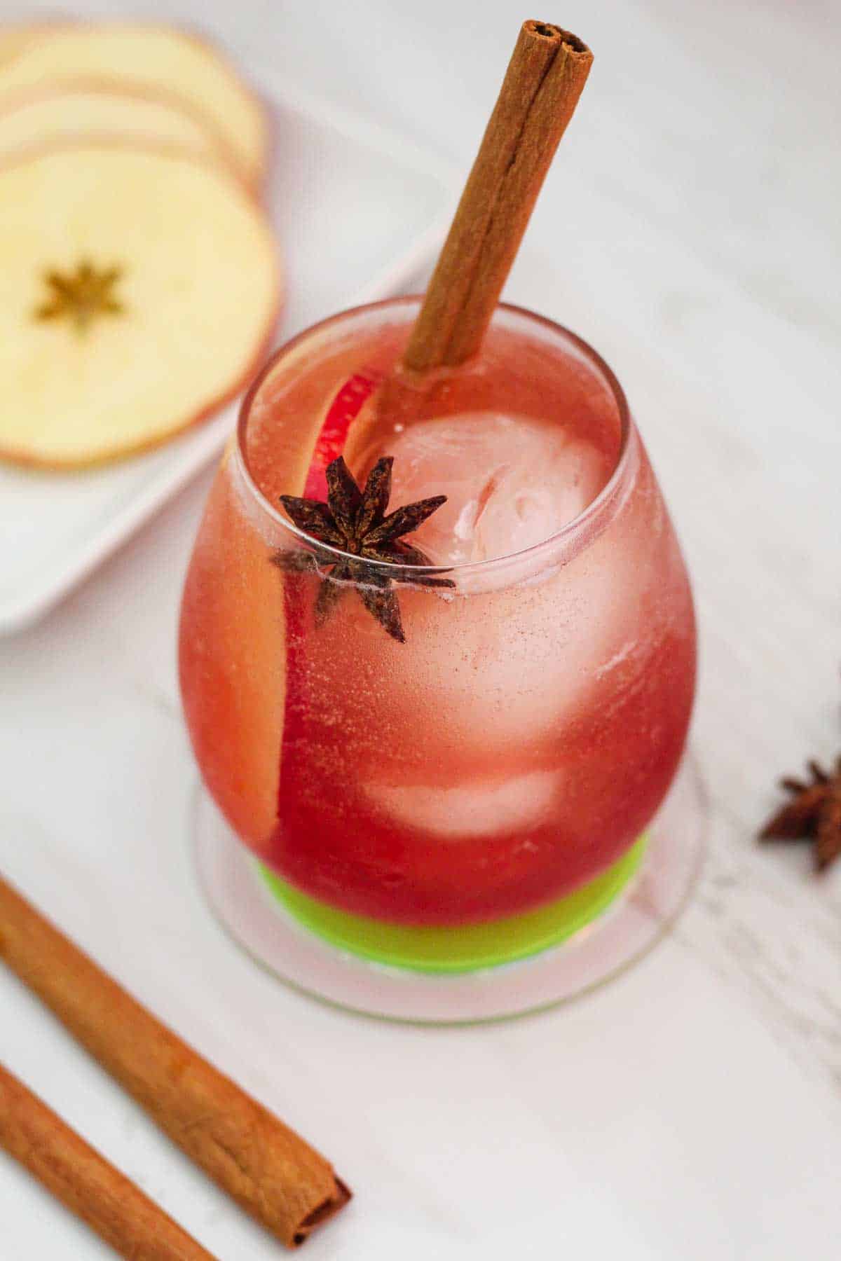 A cocktail with ice, apple slice, star anise and cinnamon stick. There are garnishes around the cocktail glass, like more apple slices, cinnamon sticks etc.