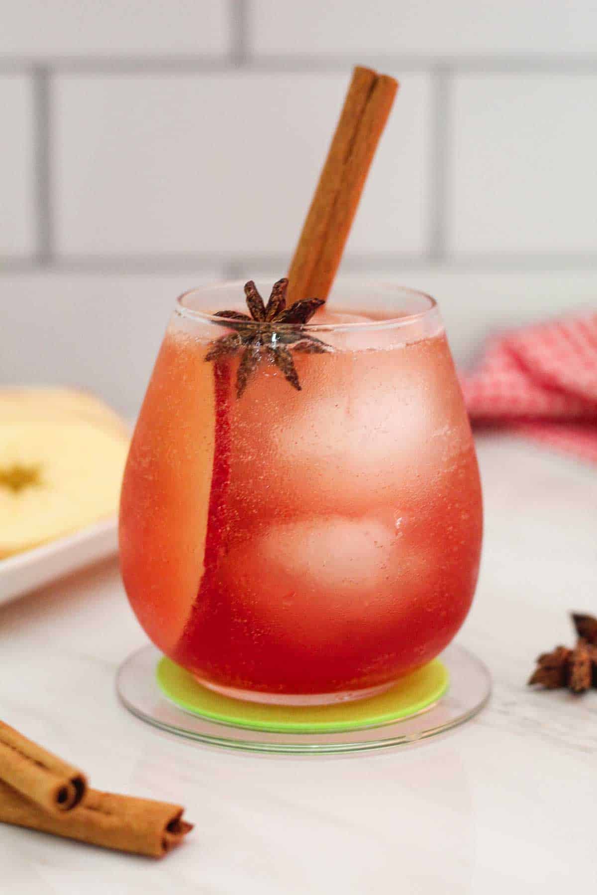 A cocktail glass with a reddish drink on ice. There's an apple slice inside the drink, a star anise and a cinnamon stick as garnishes on the drink. 