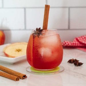 A cocktail glass with a reddish cocktail on ice. Cocktail has an apple slice, cinnamon stick and a star anise as garnishes.