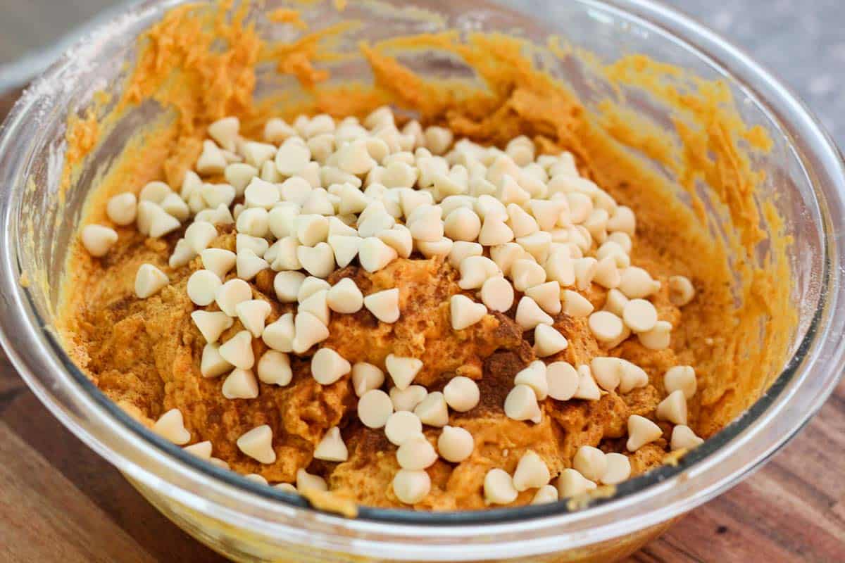 Muffins batter (looks orange from pumpkin) on a mixing bowl. On top of the batter you see seasoning and white chocolate chips that haven't been folded in yet.