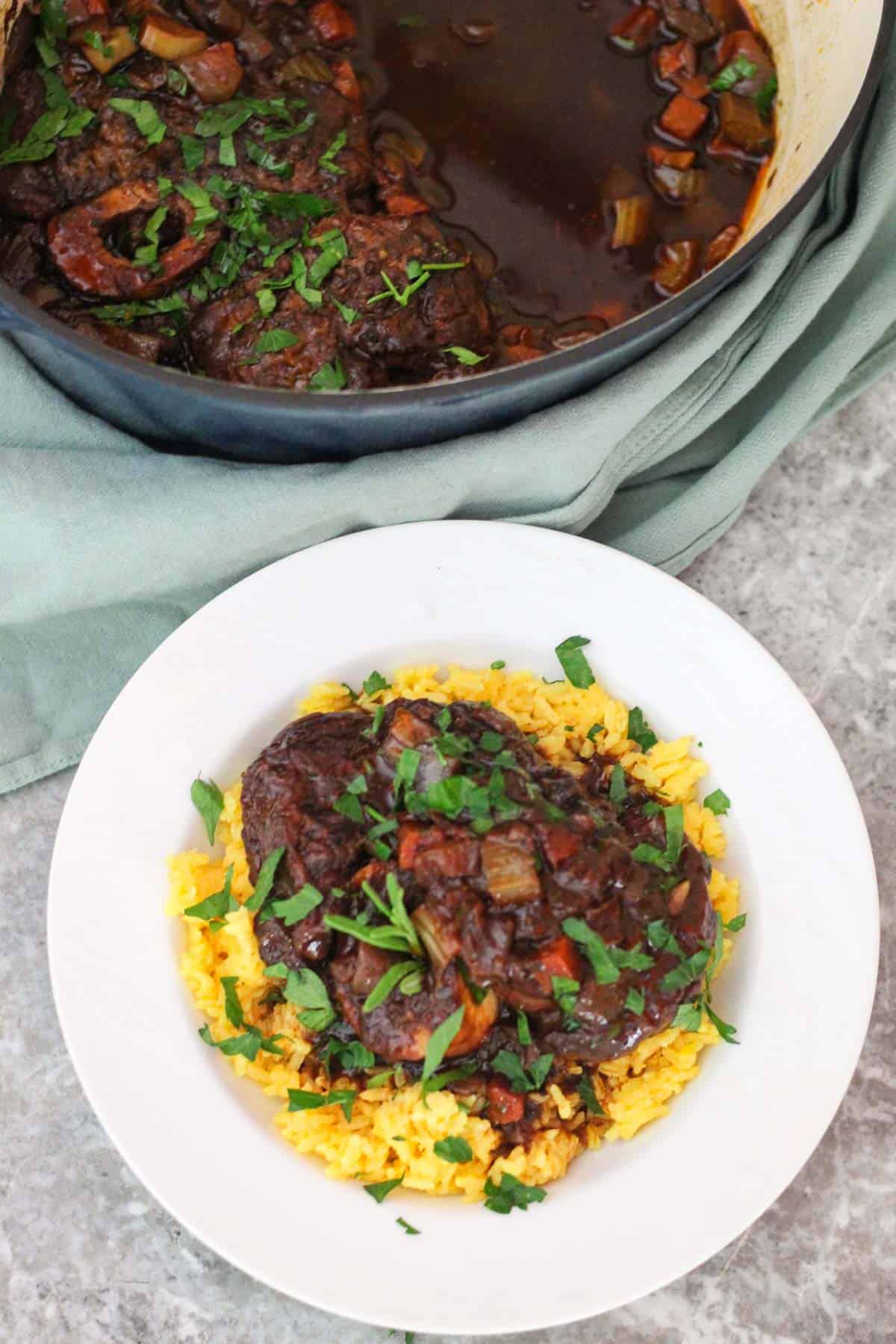 A plate with yellow rice topped with a serving of braised beef shanks. The dutch oven with the remaining beef shanks is right next to the plate.