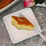 A slice of diamond shaped revani cake on a square plate, next to the tray where cake was baked. There's a rose in the background too.