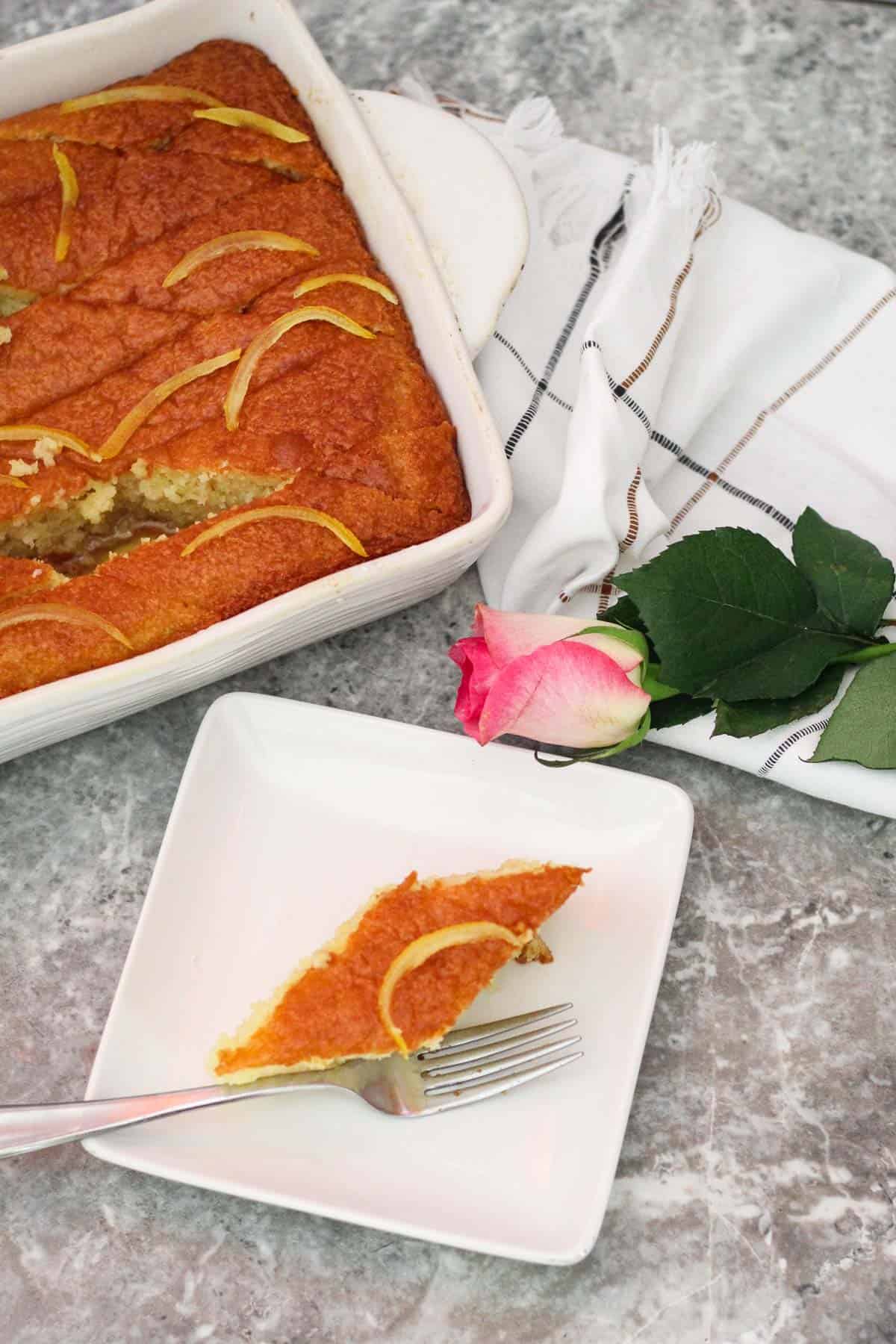A slice of revani cake on a square, white plate garnished with a lemon peel. There's a rose in the background over a kitchen towel and the revani baking dish in the background. 