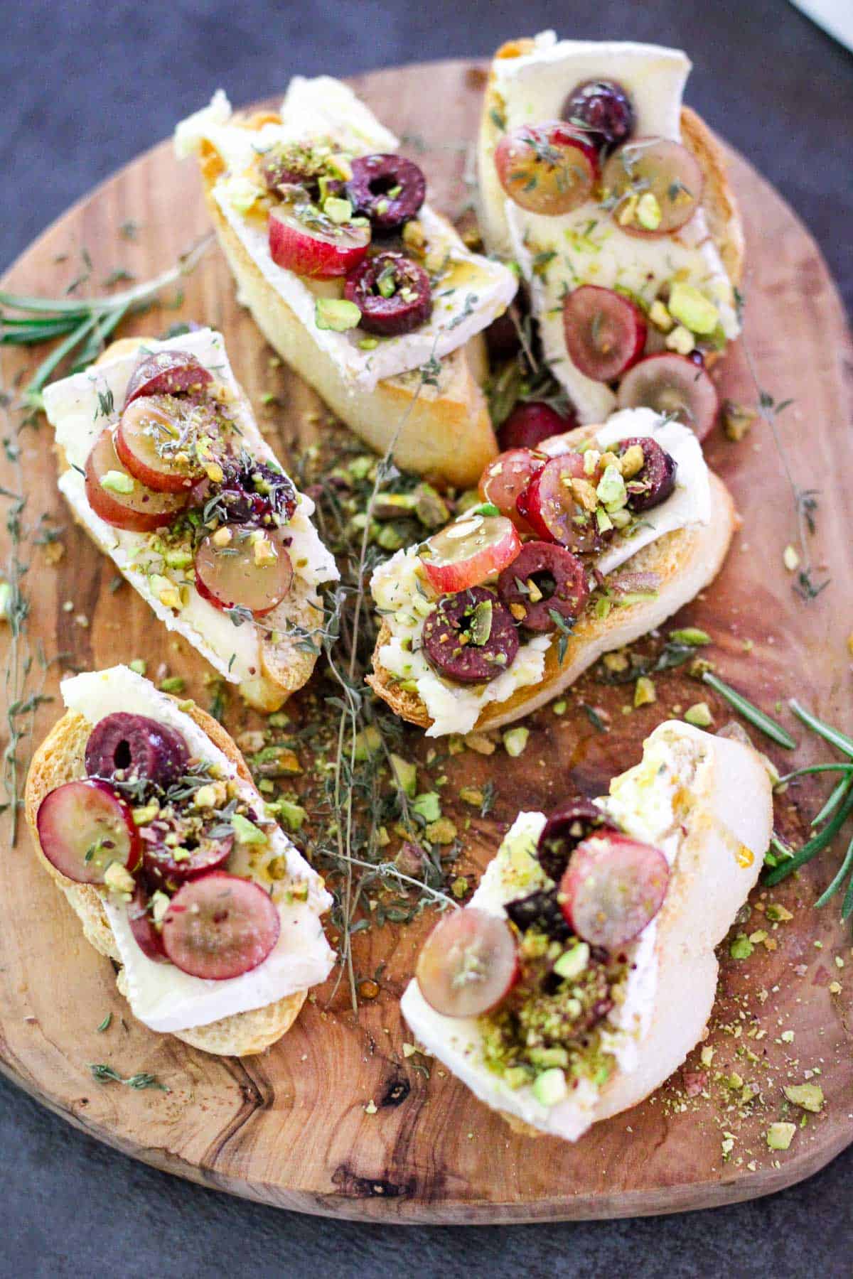 A cheese wood board shown with crostini on top. The bread bites have brie cheese, grapes, olives, pistachios visibly on them and some garnishes with thyme and rosemary.
