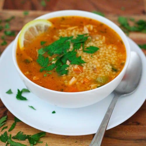 A bowl of chicken pastina soup shown with a spoon next to it. Bowl is garnished with fresh Italian parsley.