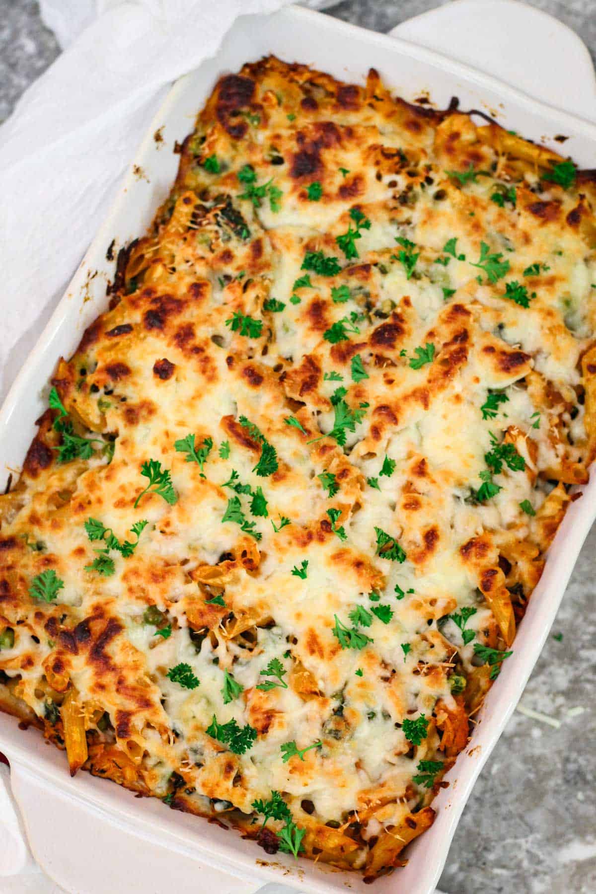 A casserole dish with baked penne pasta, covered in cheese and garnished with fresh parsley.