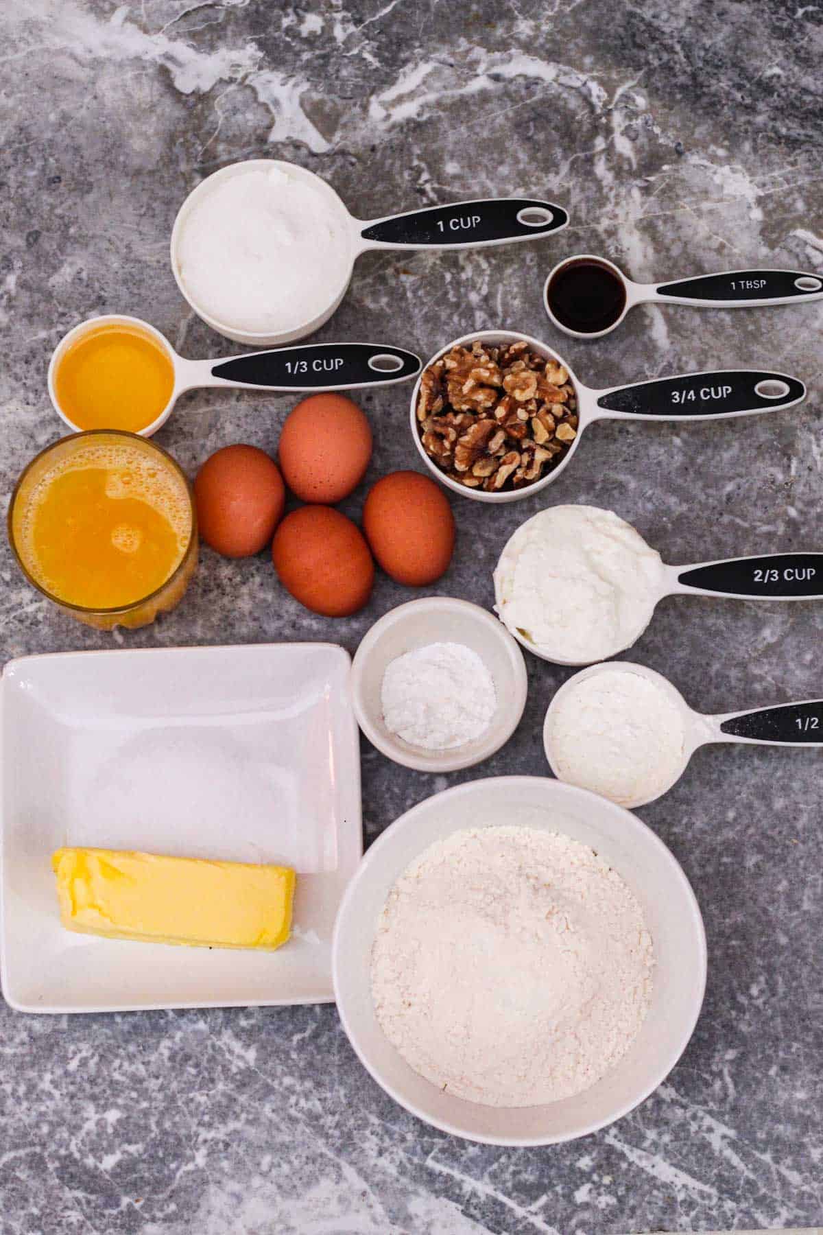 Orange bundt cake ingredients in measuring cups or spoons: Flour, butter, baking powder, cornstarch, yogurt, eggs, walnuts, orange juice, avocado oil, sugar and vanilla extract.
