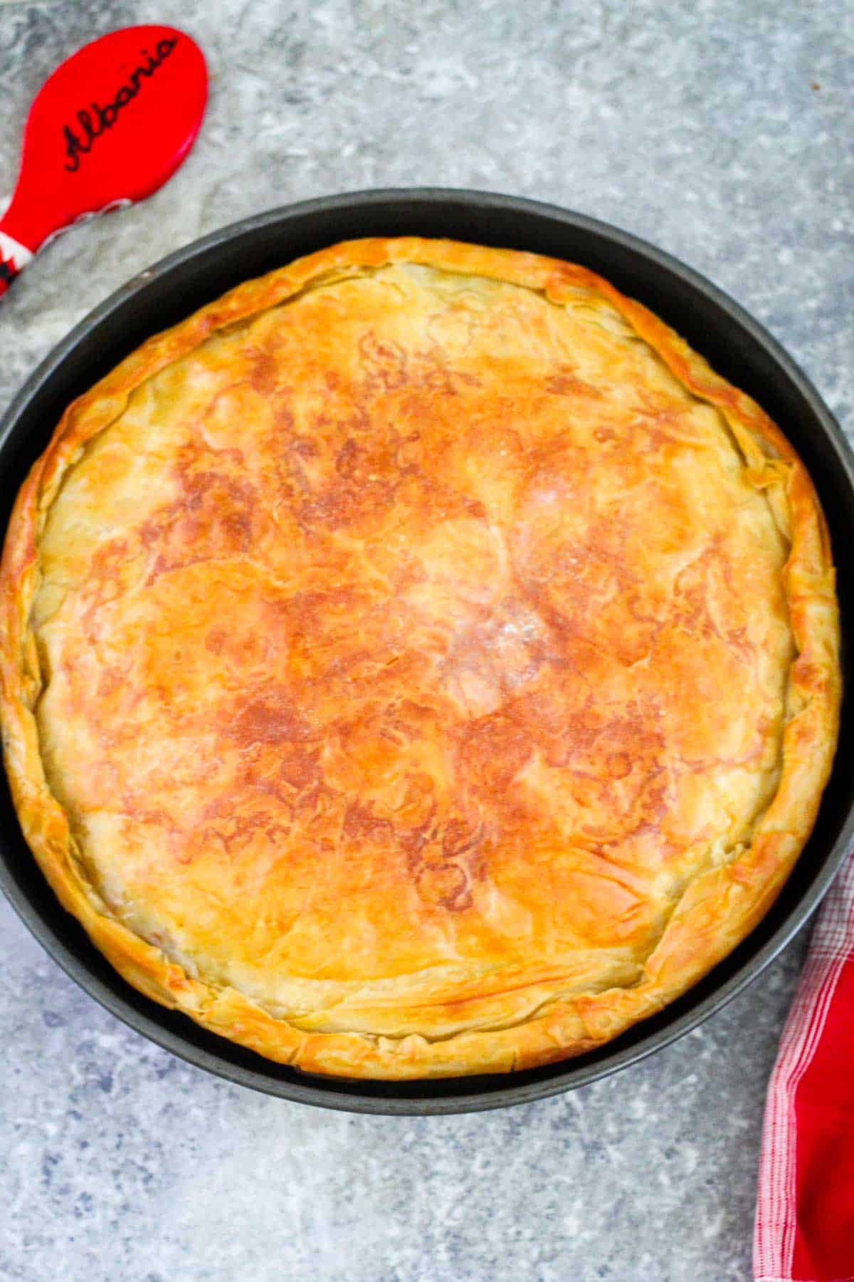 A round baking tray that shows a baked phyllo meat pie. Pie is tucked in on the edges and looks golden brown crispy. There's a red wooden spoon that reads Albania on top and a red kitchen towel on the right hand side of the baking tray.