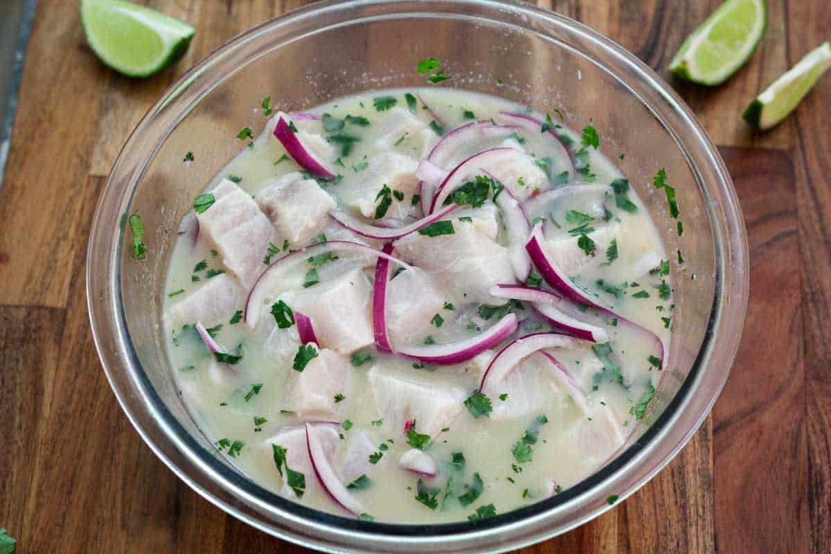 A glass bowl showing fish marinating with leche de tigre. You can also see red onions and cilantro on top of the marinate.