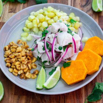 A plate with corvina ceviche, toasted corn, sweet potato slices and choclo corn, garnished with lime slices and cilantro.