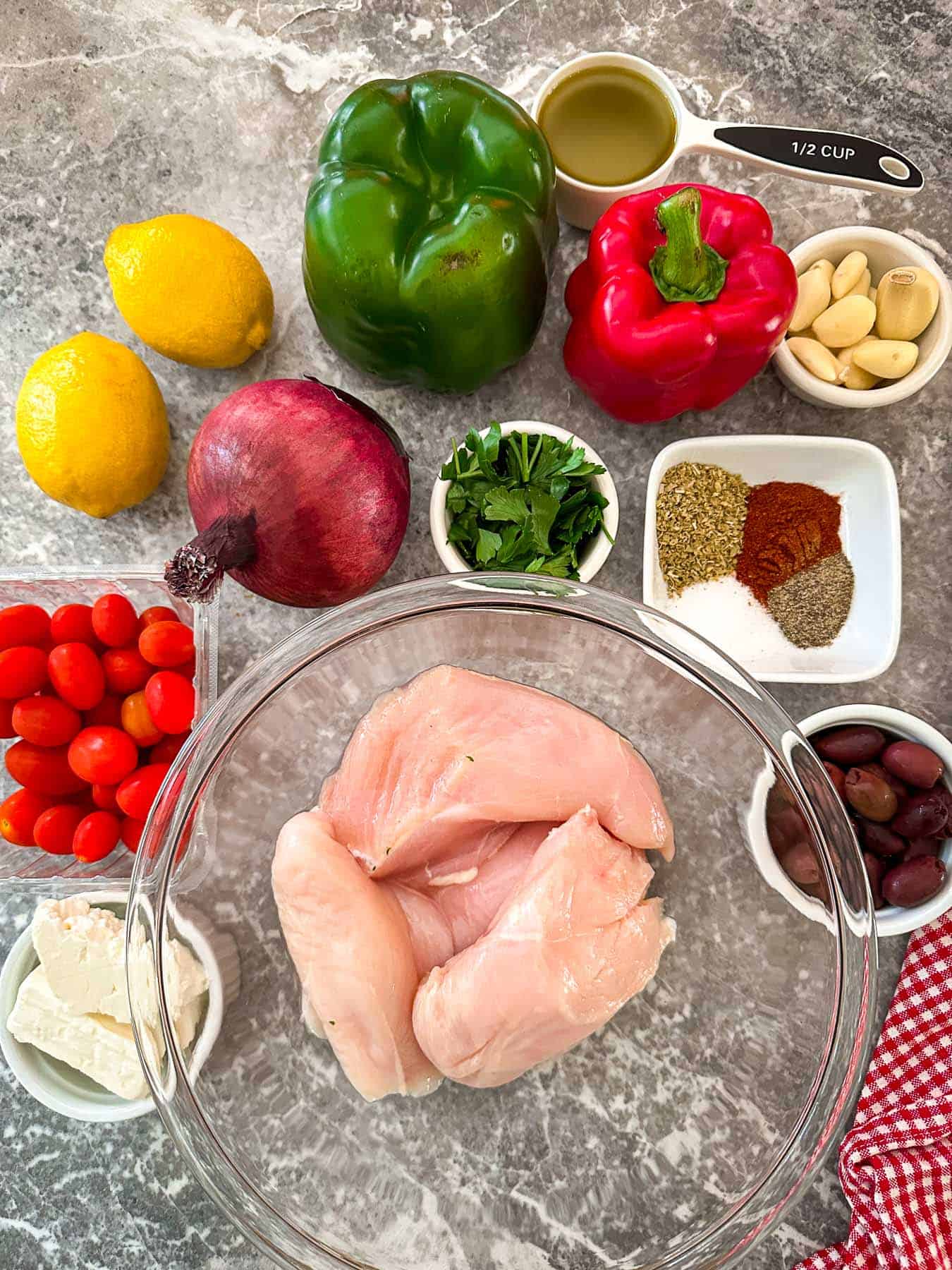 Ingredients for Mediterranean Sheet Pan Chicken dinner, laid out on a counter. You can see red onion, bell peppers, chicken, grape tomatoes, kalamata olives, feta cheese, herbs and seasonings. Also, visible are olive oil and lemons. 