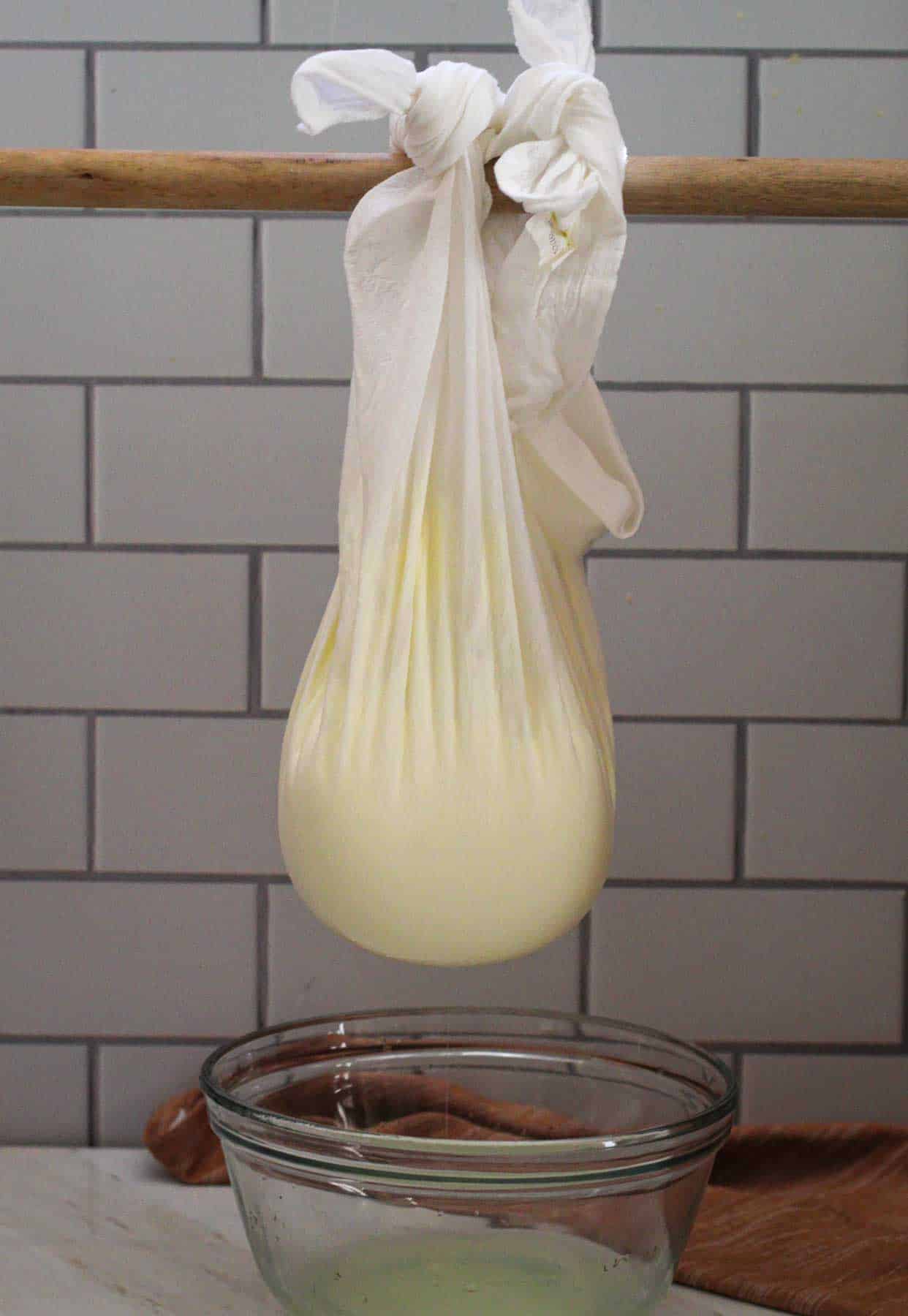 Picture shows a muslin cloth with yogurt in it, tied up and hanging from a rod. The liquid that is strained is falling over a glass container. 
