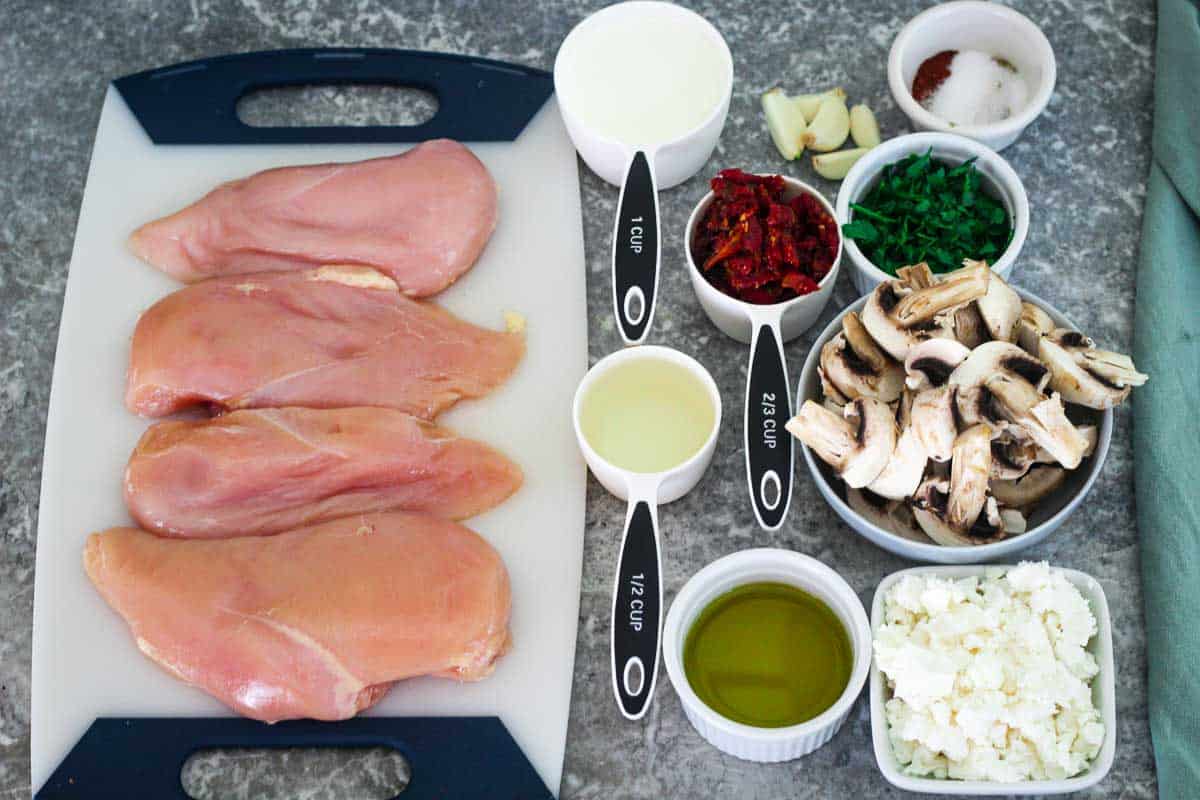 Ingredients for creamy chicken with sun dried tomatoes all grouped together before cooking: raw chicken breasts, heavy cream, white wine, olive oil, garlic, sun dried tomatoes, mushrooms, fresh parsley, salt, pepper, oregano, paprika and feta cheese, 
