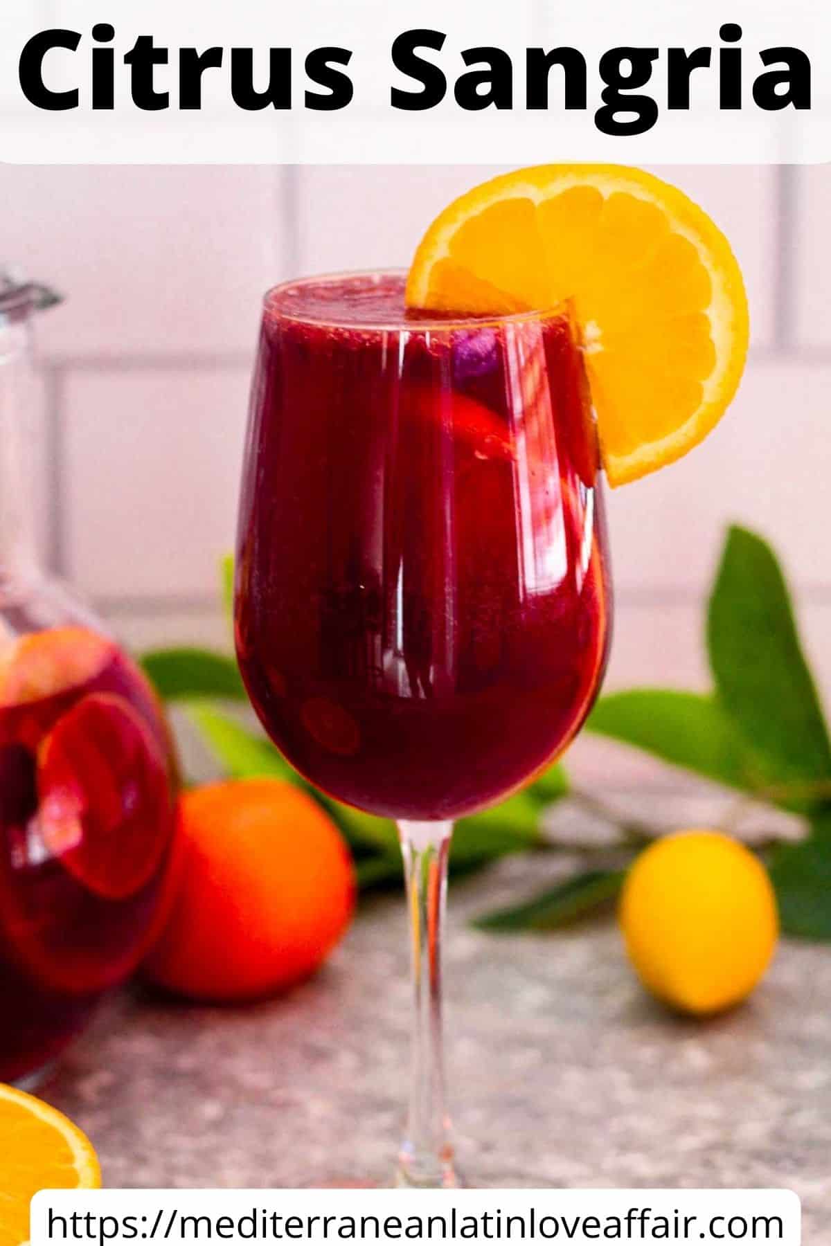 An image prepared for Pinterest. It shows a glass of citrus sangria with red wine. garnished with a slice of orange. Glass is forefront to a pitcher of sangria and more citrus fruits around the counter. 