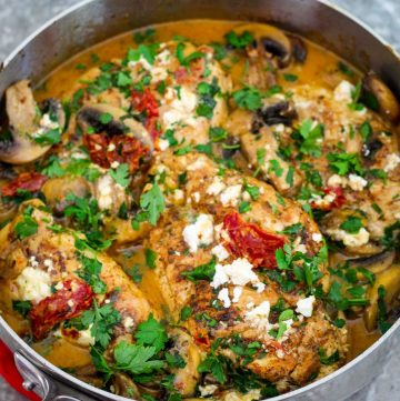 A skillet showing cooked chicken in creamy sun dried tomatoes sauce and mushrooms. Dish is garnished with feta cheese and fresh parsley.