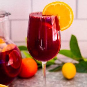 A glass of citrus sangria with red wine, shown garnished with a slice of orange. In the background you see the sangria pitcher, a mandarin, a lemon and citrus leaves.