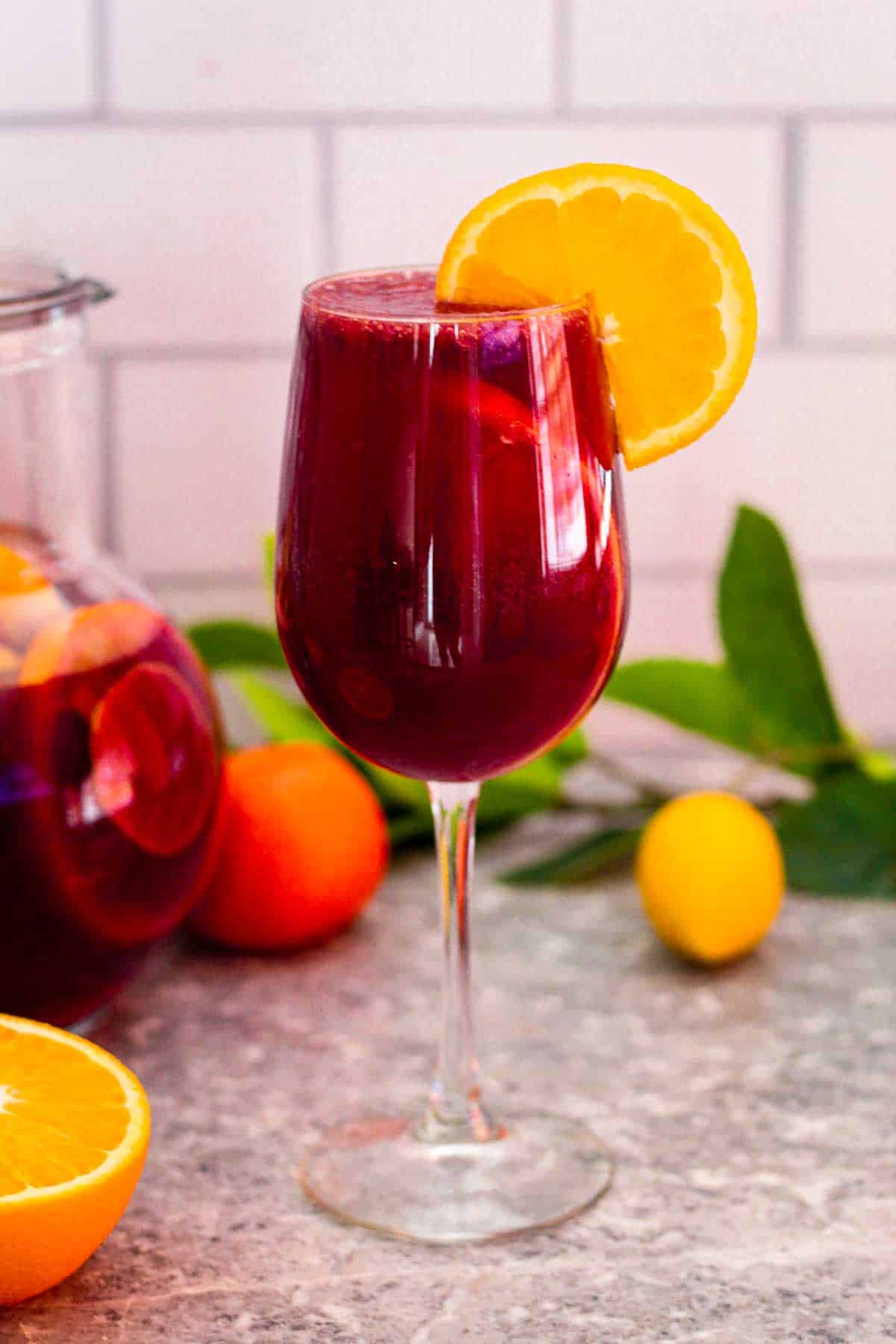 A glass of citrus sangria with red wine. Glass is garnished with a slice of navel orange. In the back