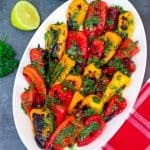 An oval platter with grilled mini peppers, topped with a greenish parsley and za'atar sauce. There's fresh parsley and lemon on the side.