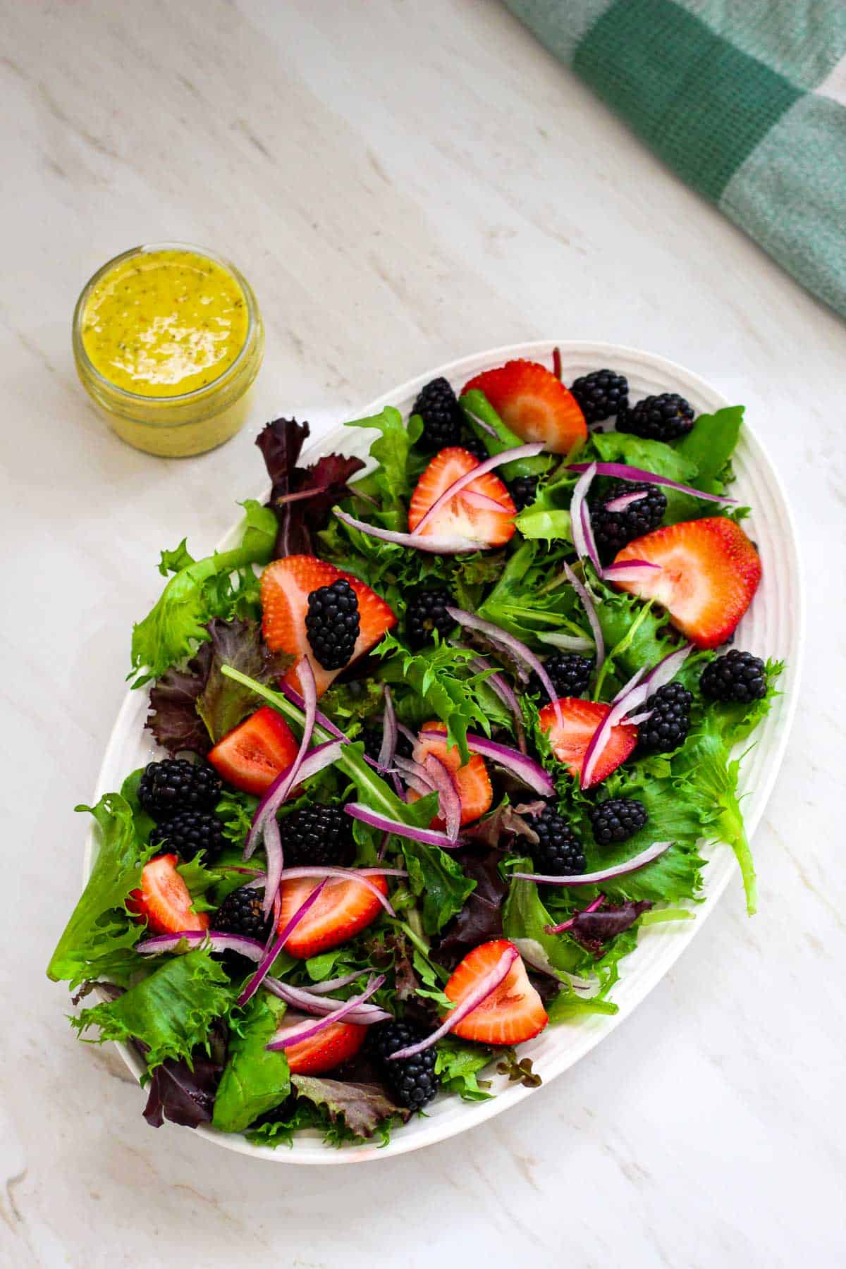 An oval platter with greens, strawberries, blackberries, onions etc. On the side of the platter you can see the vinaigrette.