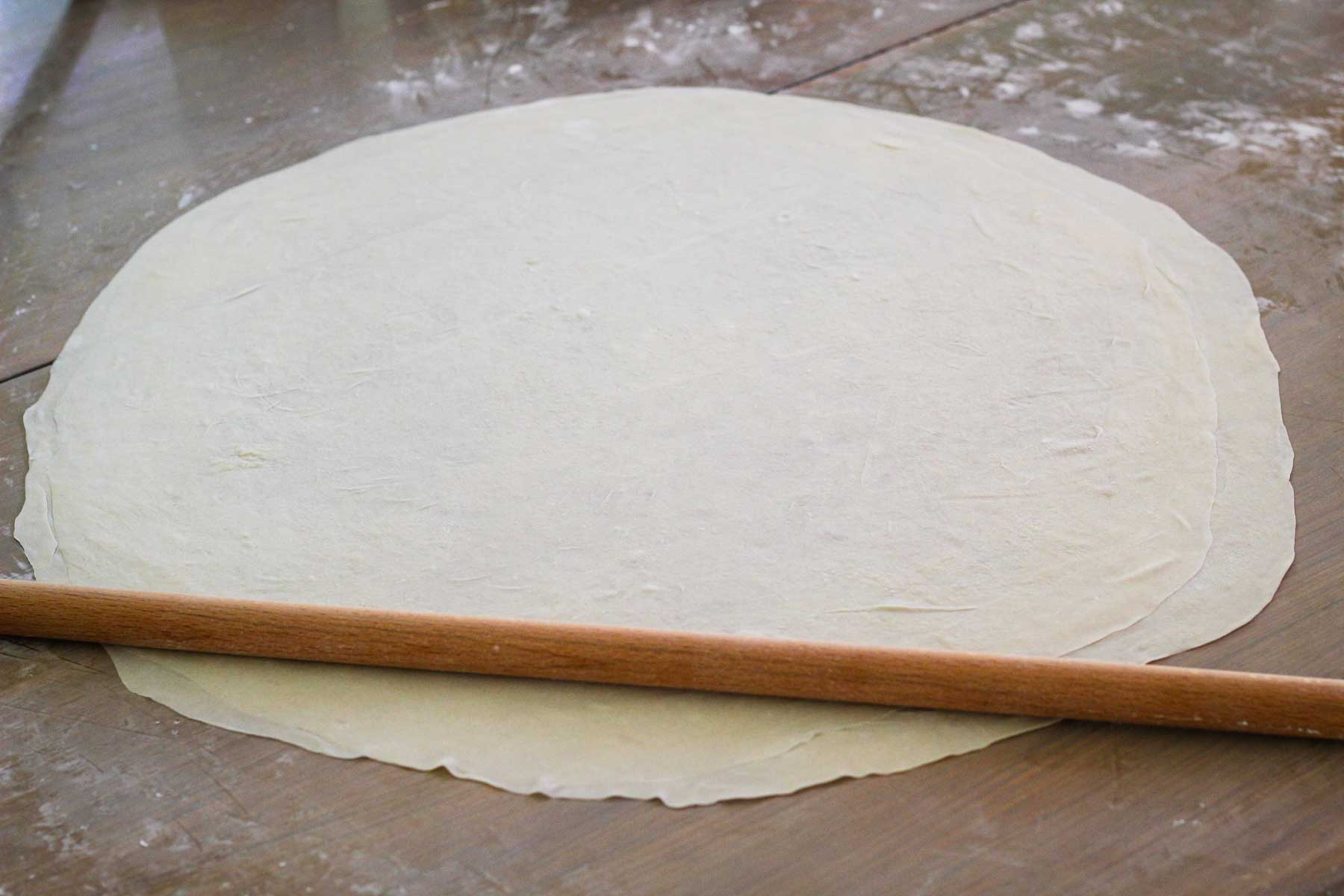 A phyllo layer stretched over the tabletop with a thin rolling pin on top.