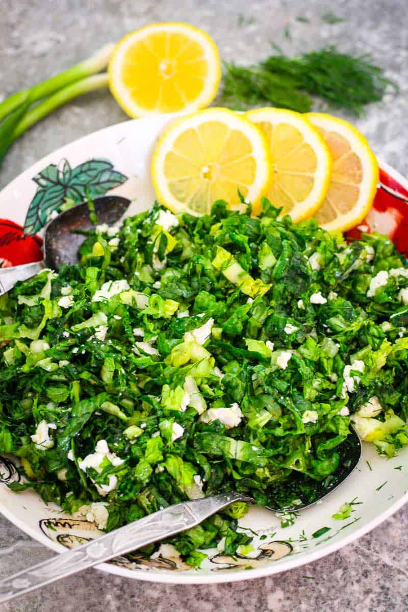Fresh lettuce salad with dill, scallions and mixed with crumbs of feta cheese. Salad is shown with fresh slices of lemon and you can see more ingredients in the background. 
