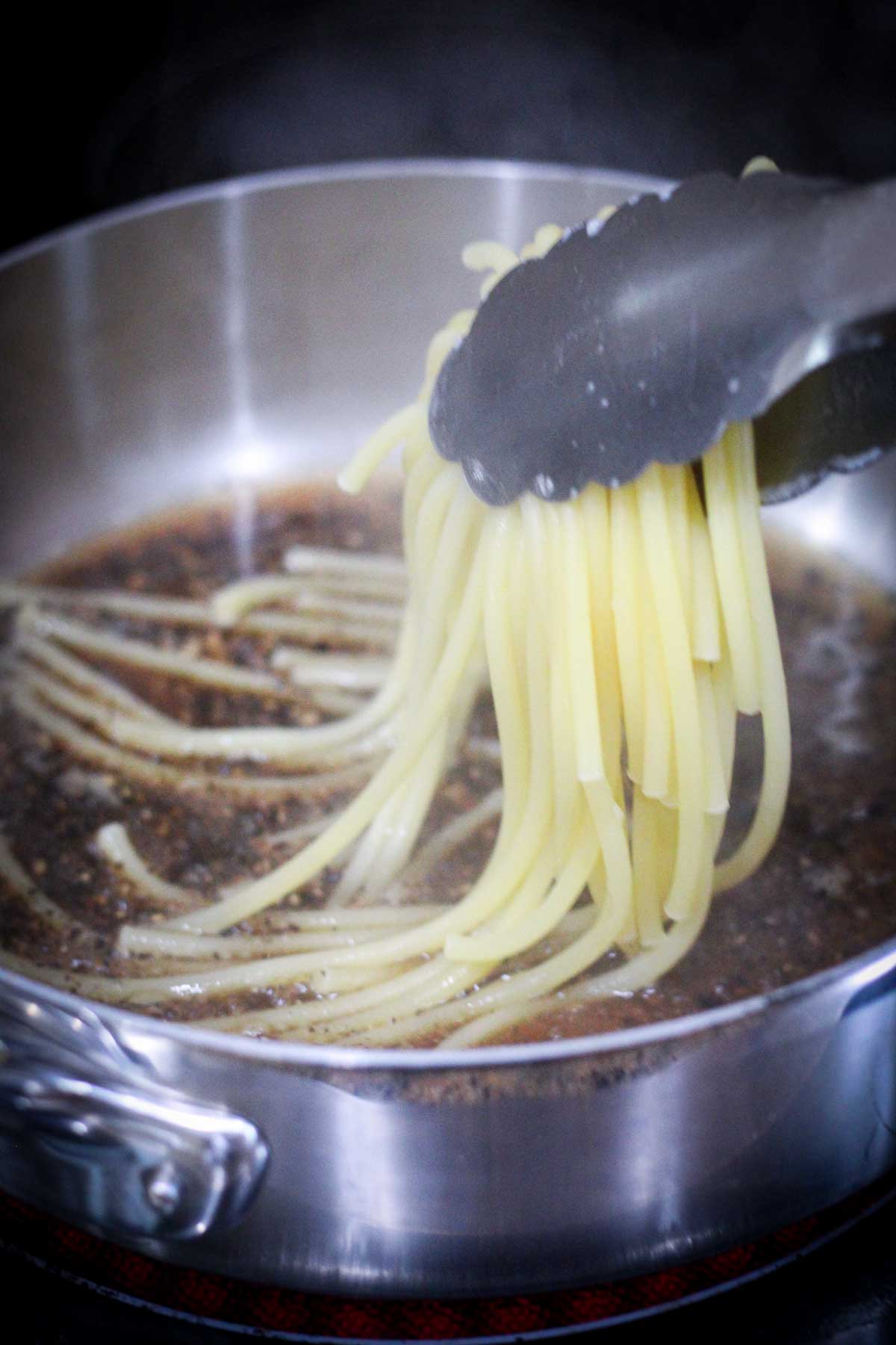 Adding pasta to peppercorn sauce in a pan using tongs.