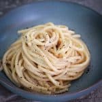 A serving of bucatini cacio e pepe on a round, bluish plate. Pasta is arranged in a nest shape.