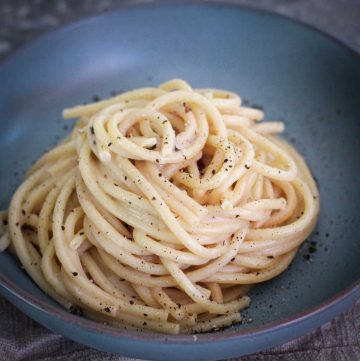A serving of bucatini cacio e pepe on a round, bluish plate. Pasta is arranged in a nest shape.